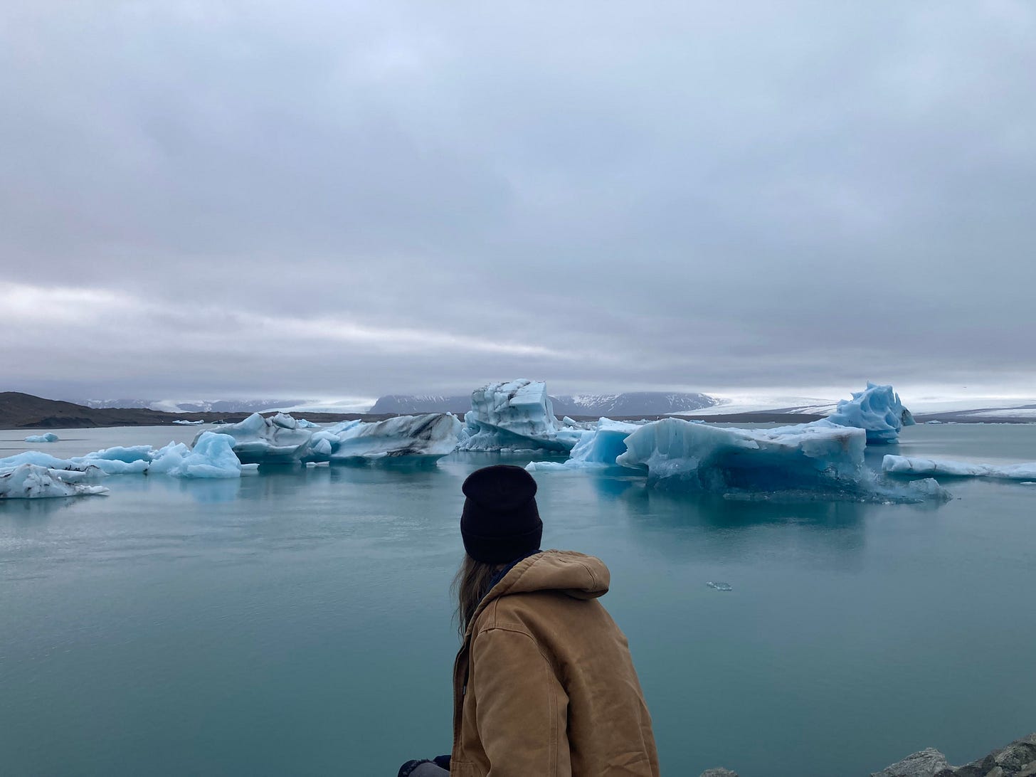 A person wearing a hat and brown jacket looks out over a glacial lagoon, with blue icebergs floating on still water beneath an overcast sky.