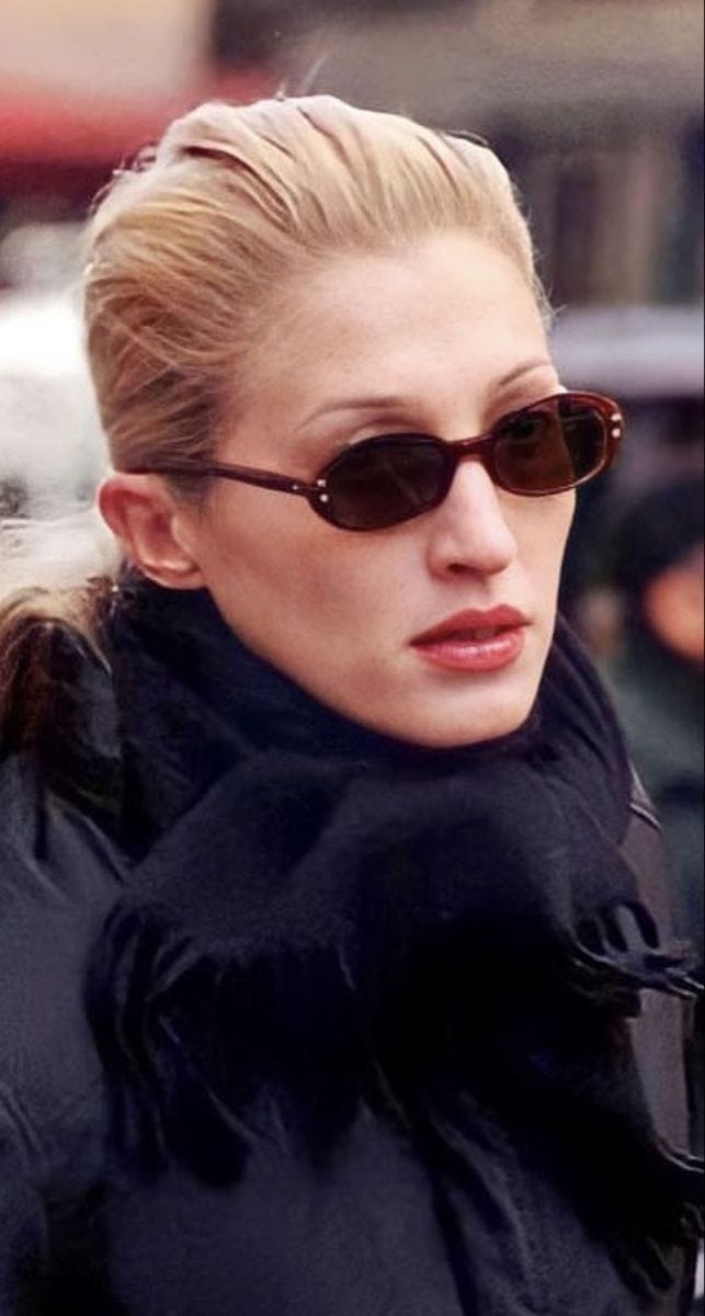 This may contain: a woman in black jacket and sunglasses walking down the street with her hand on her shoulder