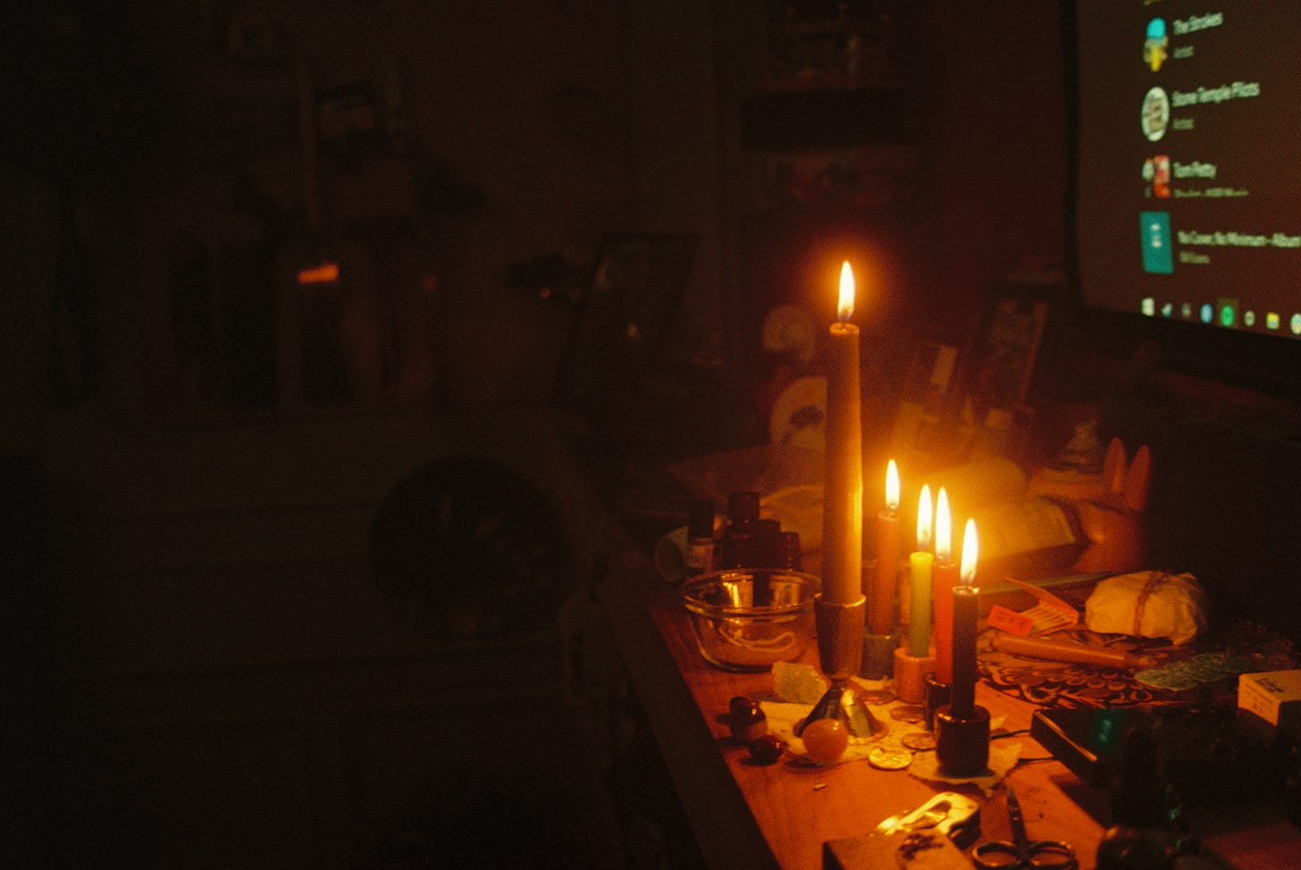 A row of four candles stands behind a larger fifth one, illuminating a wooden work table covered in various objects.
