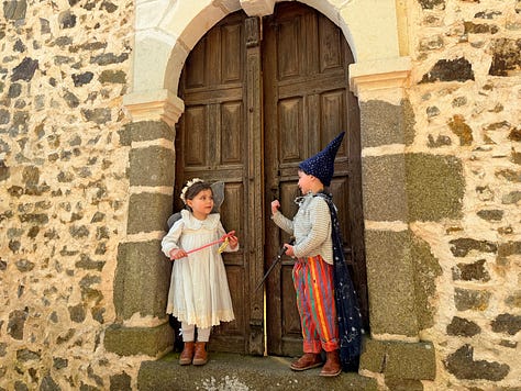 Three doors at the château: an old dark wooden door hung with a paper fairy garland, a red painted door in a stone surround with a small girl reaching for the handle, and a large arched wooden double door where two children stand dressed as a fairy and a wizard.