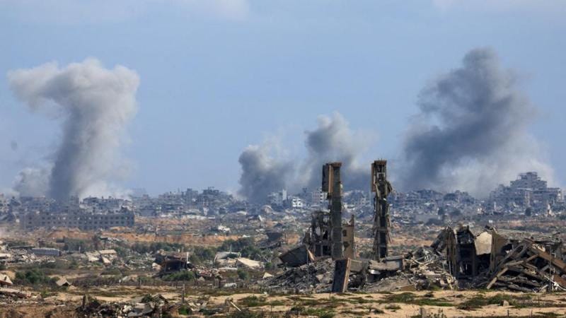 Smoke rises in the distance above the remains of lots of buildings. In the foreround, piles of debris can be seen, from what was likely buildings.