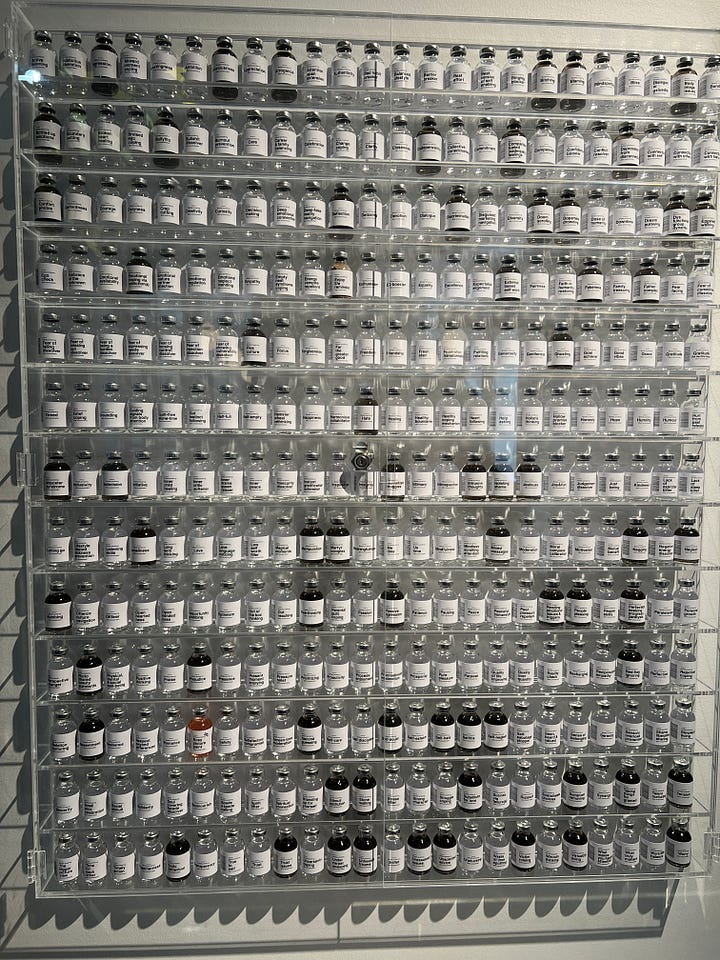 Art installation titled 'Bottled Pondering' by Aepenton, on display at the Design Museum Denmark. The piece features a wall-mounted grid of small glass vials, each labeled with a different abstract or emotional concept, such as 'Inner Critic Softener' or 'Courage Enhancer.' The vials are arranged in neat rows within a transparent display case, evoking the look of a scientific archive or apothecary cabinet of thoughts and feelings.