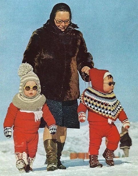 A woman in a dark coat and boots walks with two young children in red winter outfits and sunglasses across a snowy landscape. The children wear matching hats and scarves, and one has a colorful poncho. A woman in a dark coat and boots walks with two young children in red winter outfits and sunglasses across a snowy landscape. The children wear matching hats and scarves, and one has a colorful poncho.