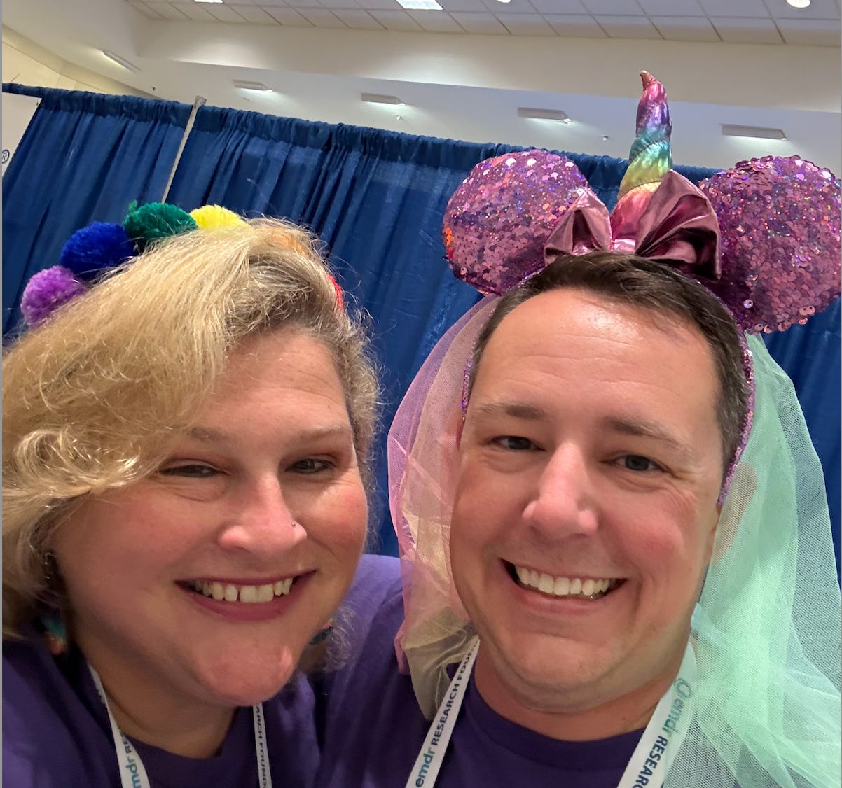 Jamie stands in a rainbow pom pom crown next to a white man weating a Minnie Mouse unicorn crown with a veil Jamie stands in a rainbow pom pom crown next to a white man weating a Minnie Mouse unicorn crown with a veil
