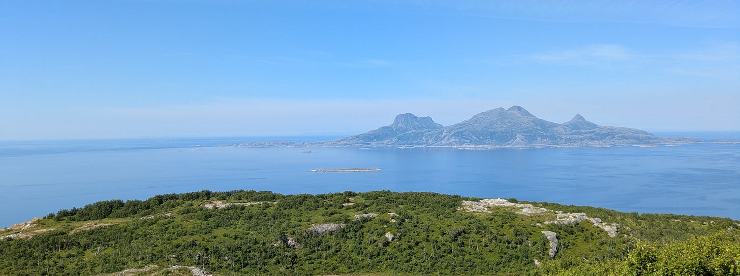 A photo from the top of a green plateau towards a mountaineous island in the sea. The water and the sky are blue