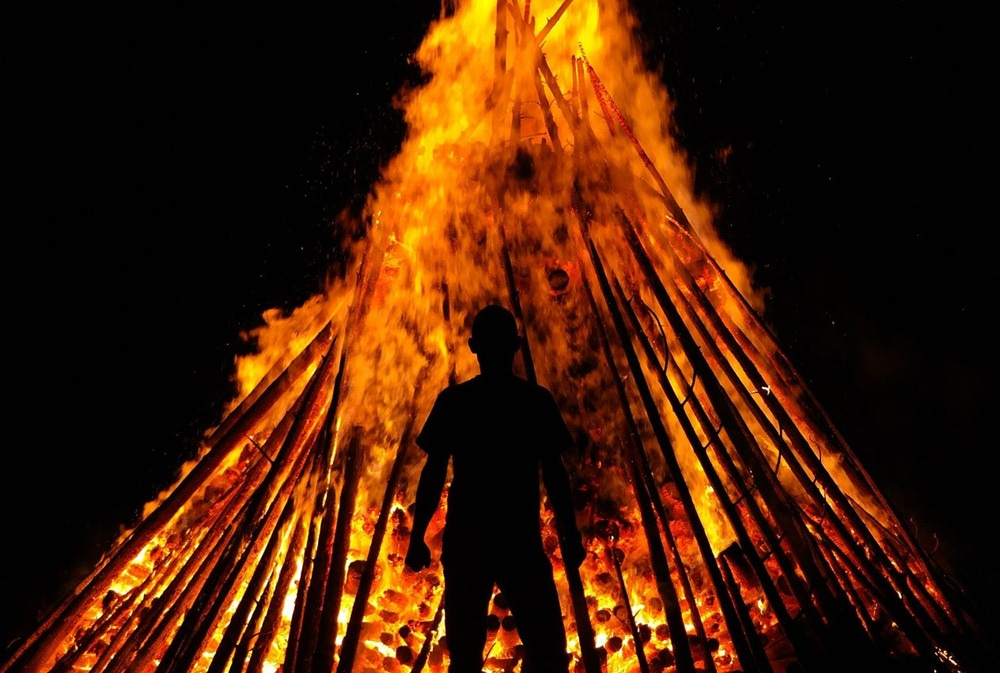A man in silhouette stands before a burning pyre.