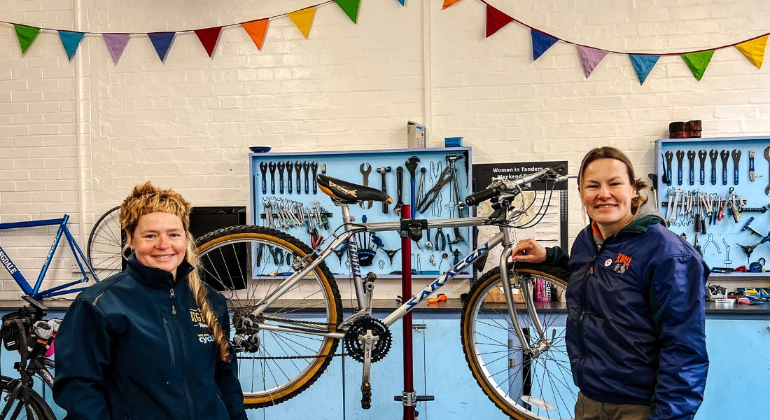 Two women standing either side of a bike, with blue cabinets, tools, and rainbow bunting behind them Two women standing either side of a bike, with blue cabinets, tools, and rainbow bunting behind them