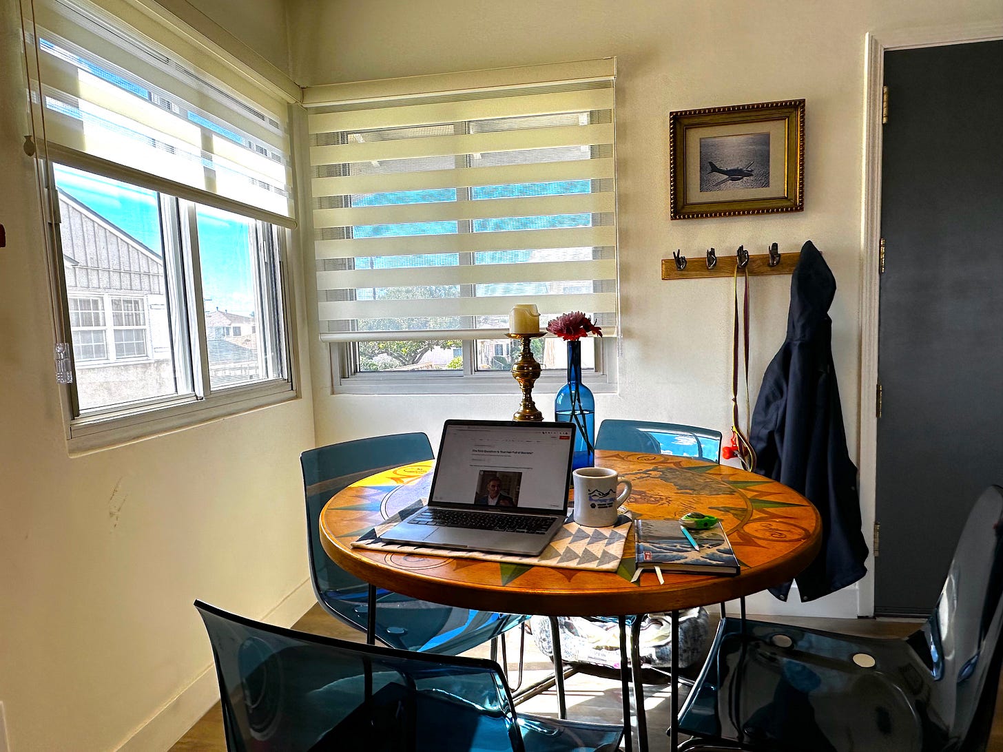 Jonathan's dining area, the relevant part of which has a circular table surrounded by 4 blue chairs. On the table is a placemat with an open laptop and cup of tea on it, and a pencil and journal to the side. Behind is a gold candlestick and blue glass bottle with some flowers in it.