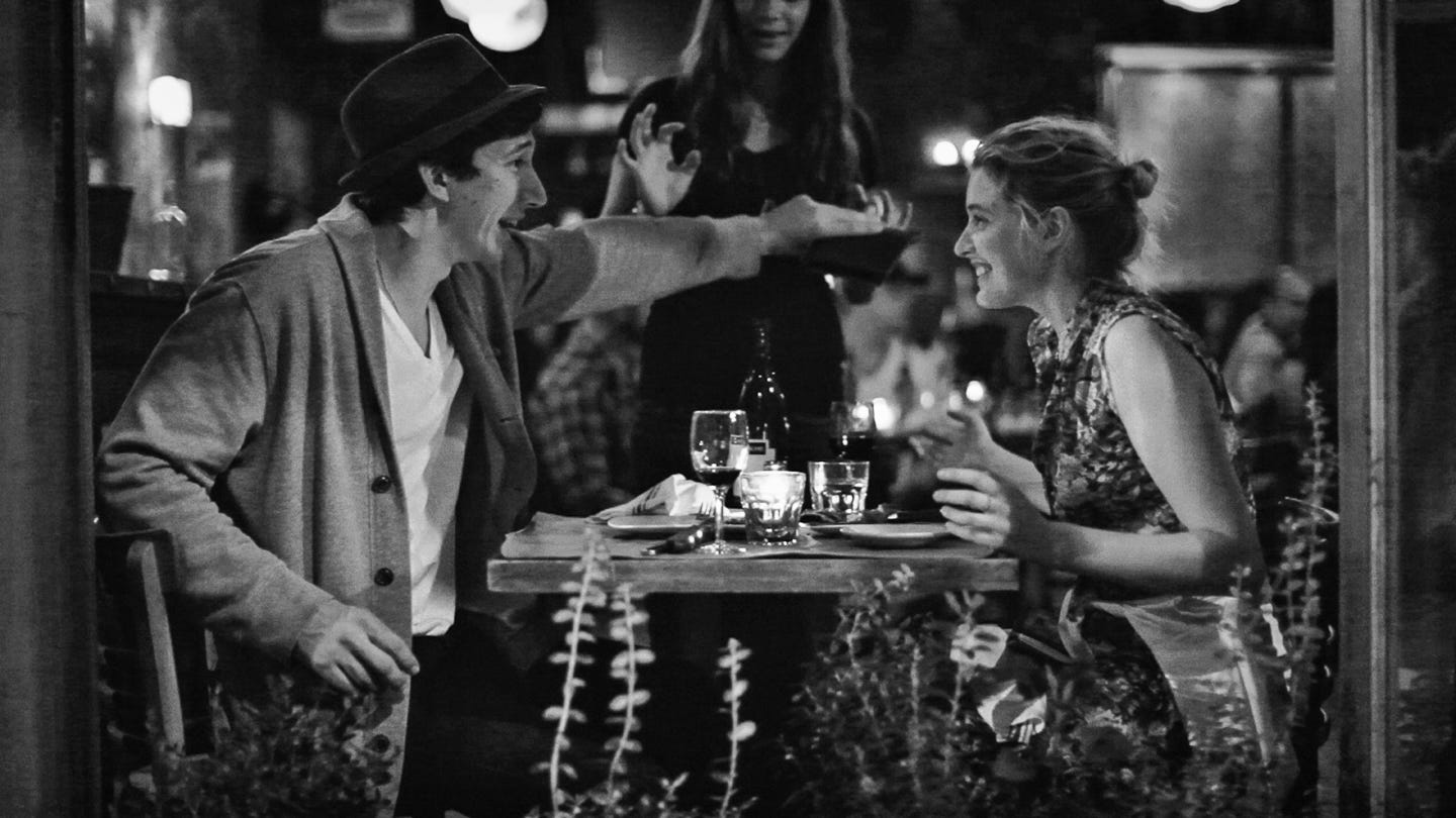 A black-and-white photo of a candlelit dinner scene. Greta Gerwig smiles brightly across the table at Adam Driver, who reaches out mid-laugh. A waitress stands between them, mid-gesture. The table is cluttered with glasses and soft light, evoking a fleeting moment of connection. A black-and-white photo of a candlelit dinner scene. Greta Gerwig smiles brightly across the table at Adam Driver, who reaches out mid-laugh. A waitress stands between them, mid-gesture. The table is cluttered with glasses and soft light, evoking a fleeting moment of connection.