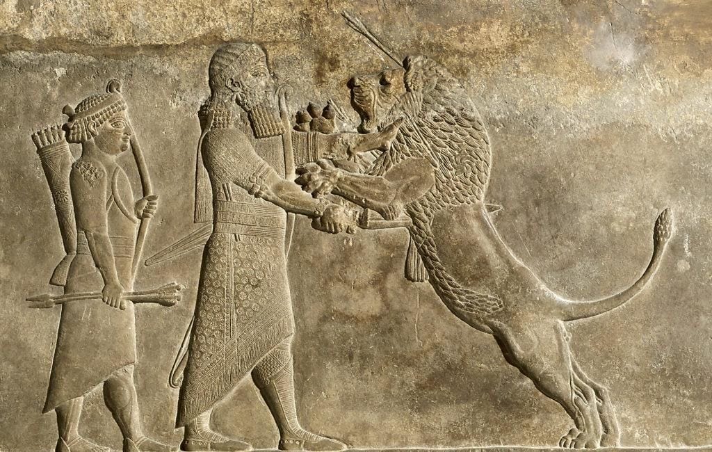 A lion which has been shot in the head with an arrow jumps at Ashurbanipal. An archer stands behind.