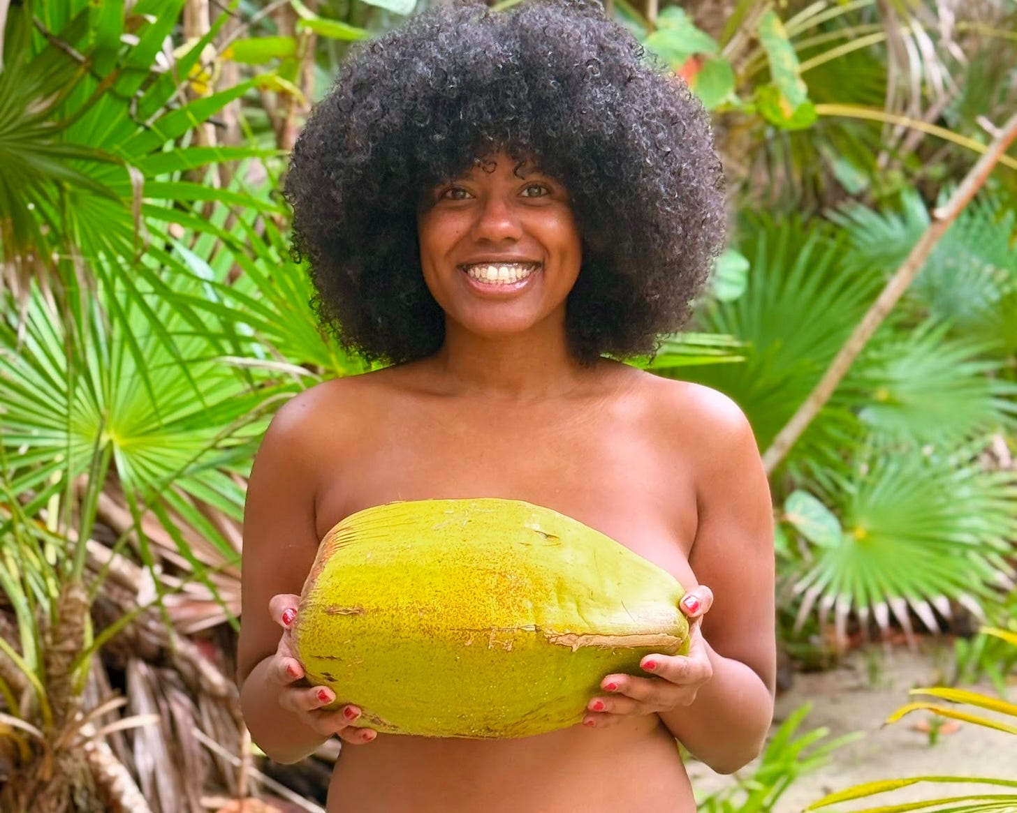 Gabriella Quinn, known as The Crowned Nudi, smiling while standing topless in a tropical setting, holding a large green coconut in both hands, surrounded by palm leaves at Playa Sonrisa in Costa Maya, Mexico.