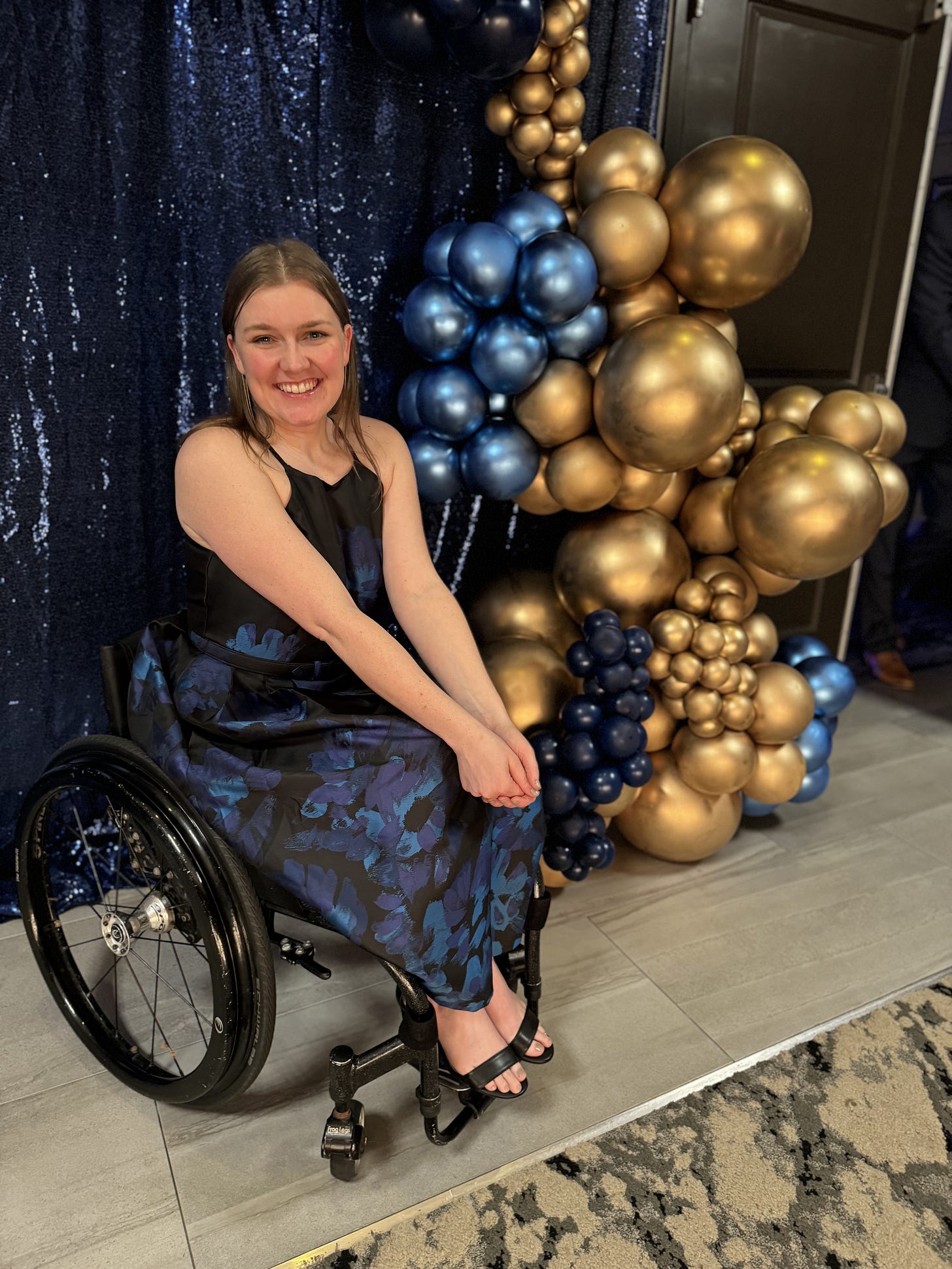 Meredith, a wheelchair user in a formal floral gown posing infront of a blue sequin background and decorative gold ballons. 