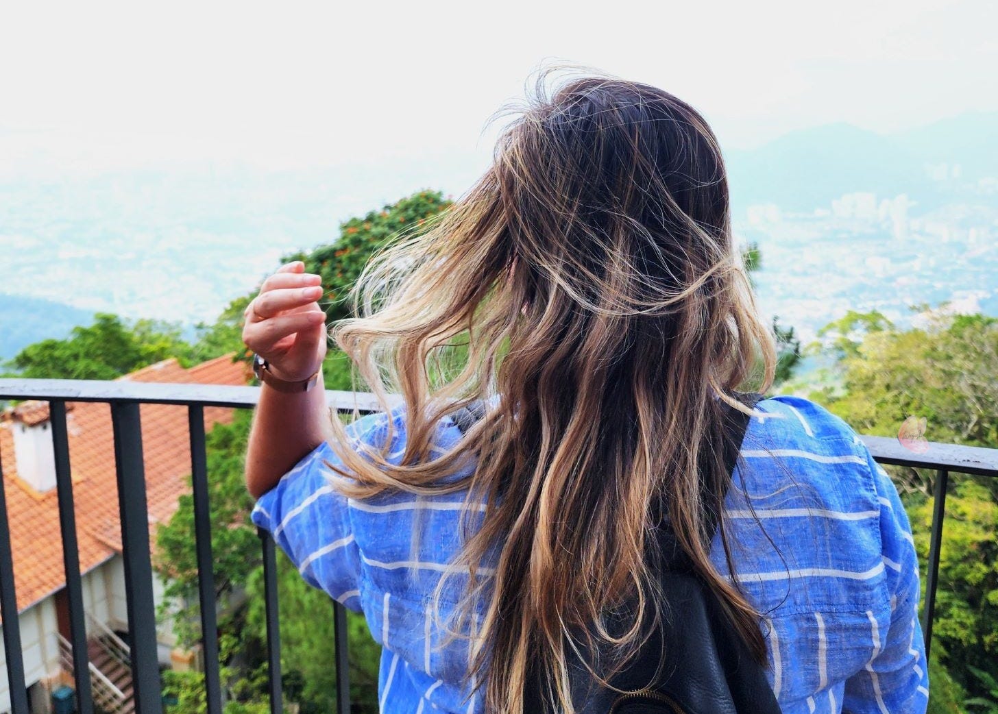 Ann standing with her back to the camera, overlooking the lush landscape of Penang Hill. Her hair gently blows in the wind, and she’s wearing a blue striped shirt.