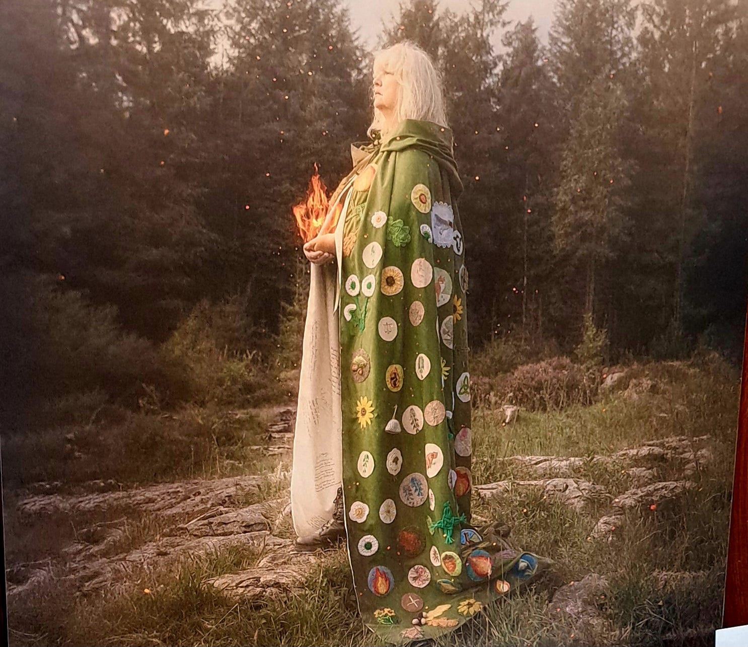 Woman with long blonde hair in profile carrying a flame in both hands at her waist. She is wearing the long green decorated cloak which pools on the grass at her feet. Behind her are tall trees, and the ground is a patchwork of rock and grass.