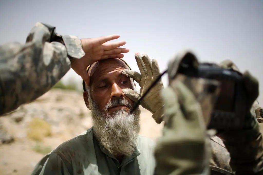 A close-up outdoor scene shows a bearded man shielding his eyes while two soldiers in camouflage and gloves examine his face, with one soldier holding a piece of equipment in the foreground against a bright, arid landscape. A close-up outdoor scene shows a bearded man shielding his eyes while two soldiers in camouflage and gloves examine his face, with one soldier holding a piece of equipment in the foreground against a bright, arid landscape.