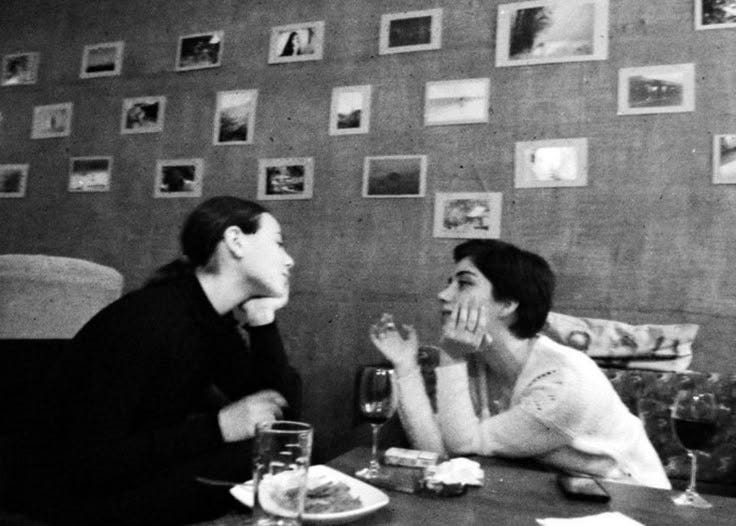This may contain: two people sitting at a table with wine glasses in front of them and pictures on the wall behind them