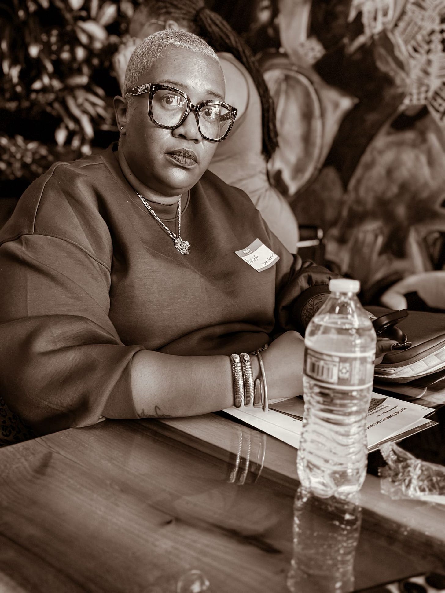 picture of a black woman with blonde, short hair wearing wide frame glasses sitting at a table. She is wearing a sweatshirt with gold bangles. On the table is a notebook, bottle of water, and other miscellaneous things. She has an inquisitive but blank stare on her face, almost as if she didn’t expect her picture to be taken. Her name is Alisha Gordon.