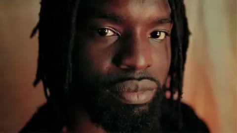 Meji Alabi A close-up of Meji Alabi, with beard and dreads, looking into the camera