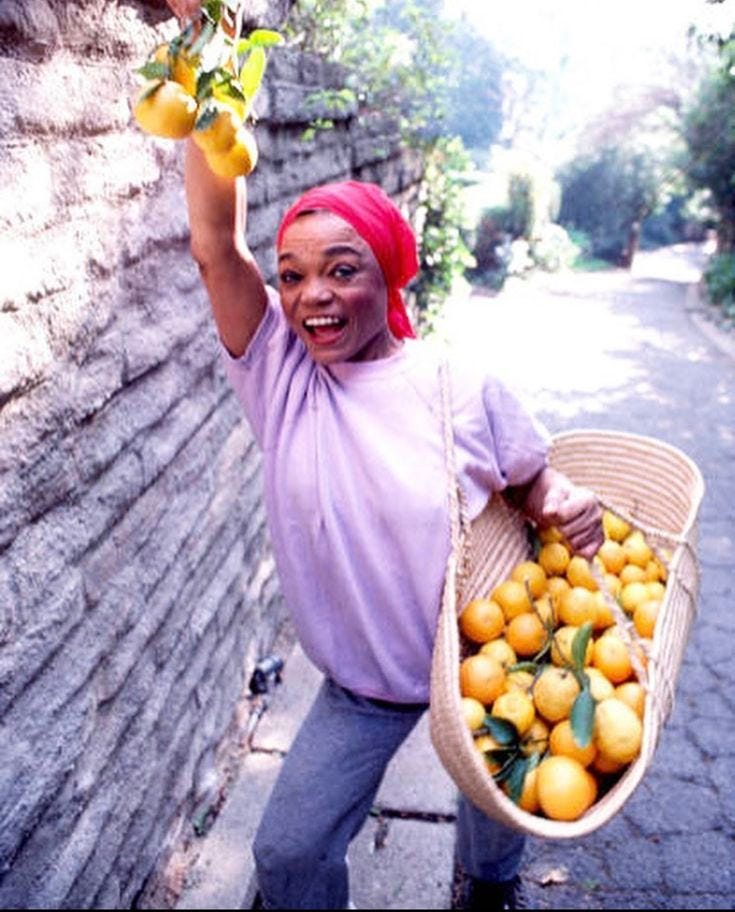 This may contain: a woman holding a basket full of oranges on the side of a road in front of a brick wall