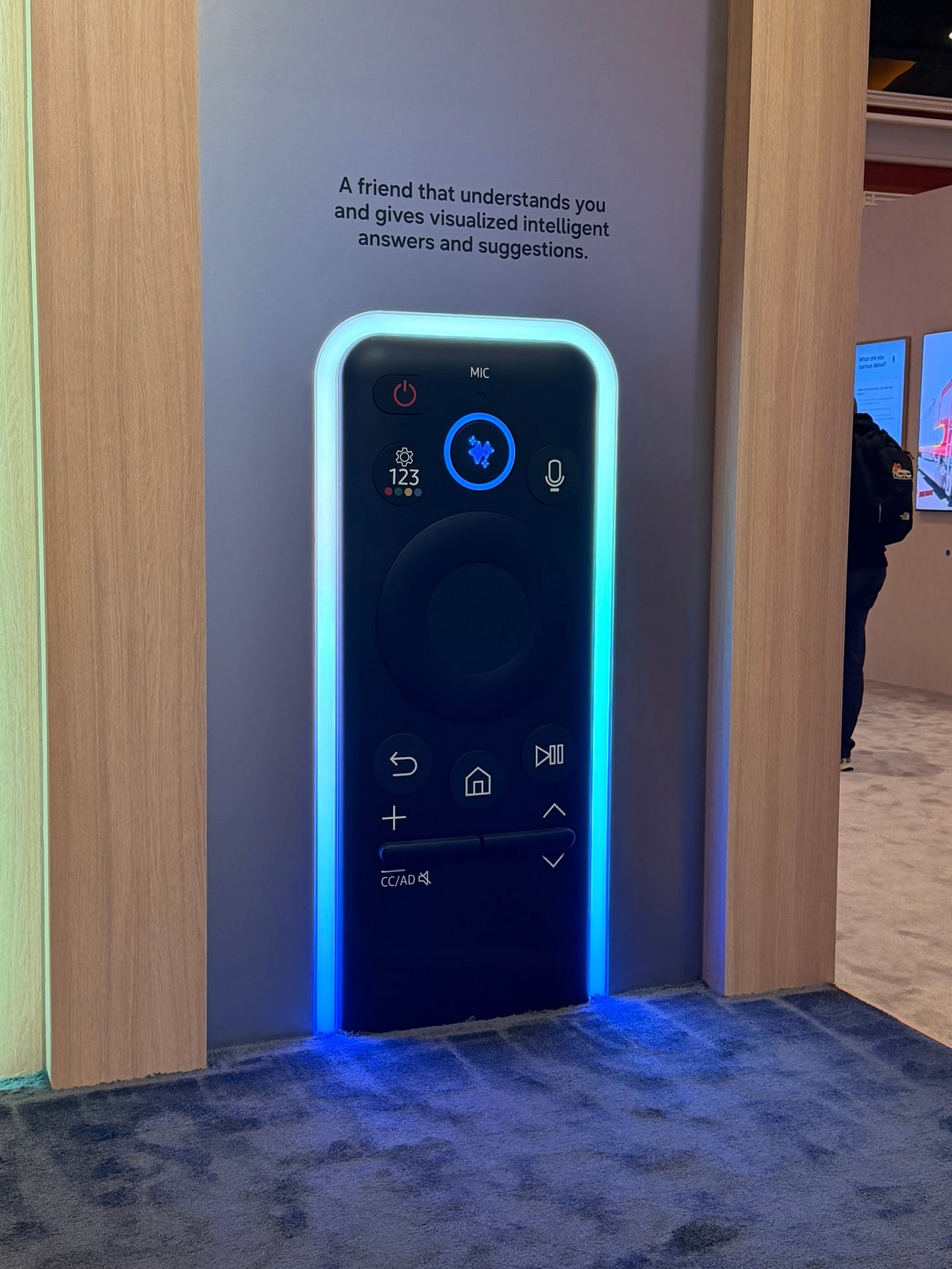 A large wall installation shaped like an oversized Samsung remote control glows with blue lighting, accompanied by text describing it as a friend that provides intelligent visualized answers and suggestions. A large wall installation shaped like an oversized Samsung remote control glows with blue lighting, accompanied by text describing it as a friend that provides intelligent visualized answers and suggestions.