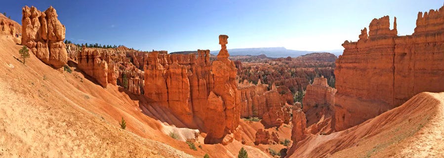 Photo taken at Bryce Canyon National Park. The immediate foreground shows sandy slopes converging and leading to unusual tall spires of rock. The orange chimneys wear in interesting patterns leaving bands of lighter and darker orange.