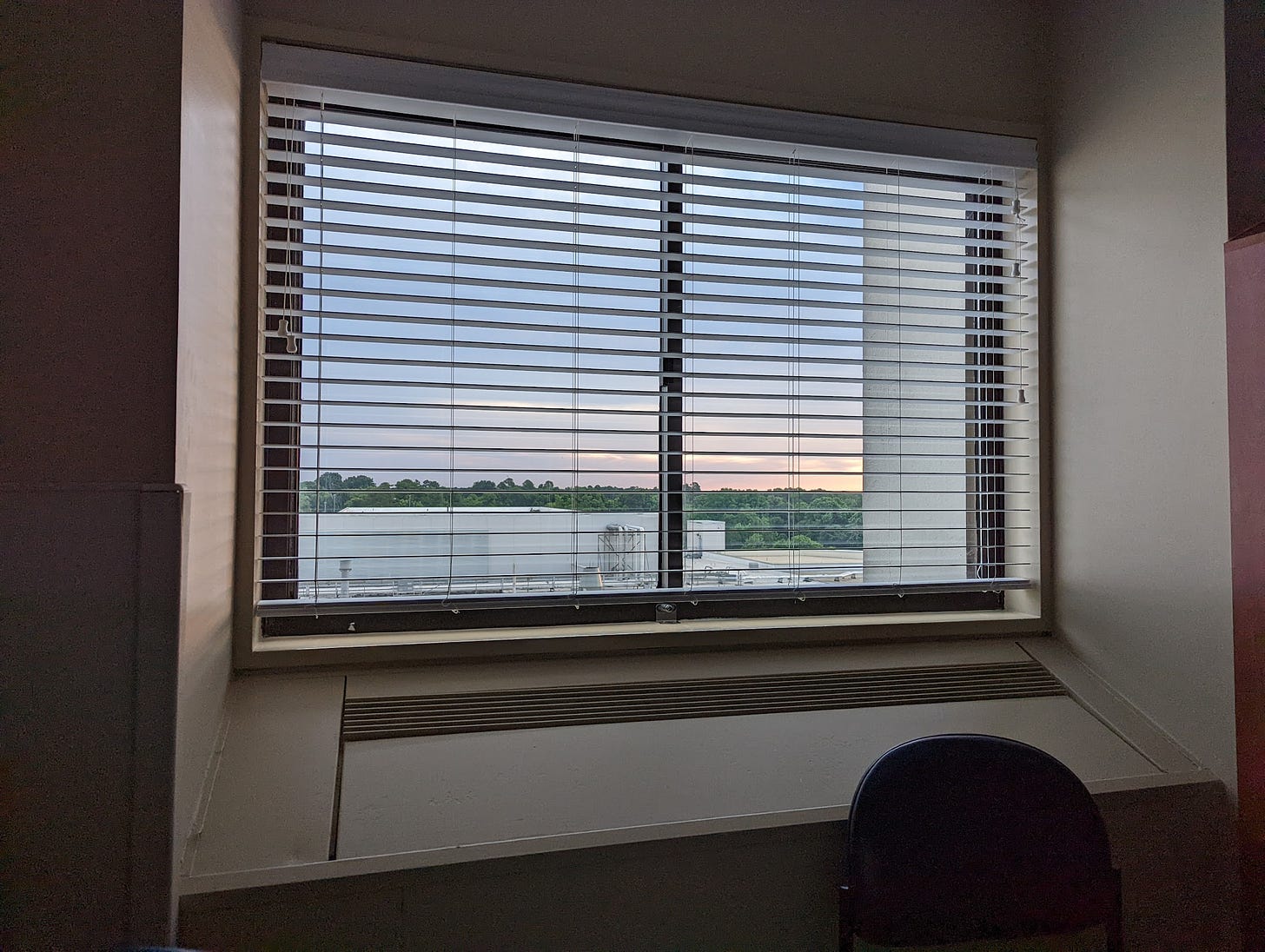 A window with blinds and a built-in AC unit. The view is of industrial HVAC equipment on a roof painted white, a tree line in the distance, and the sun rising through some clouds.