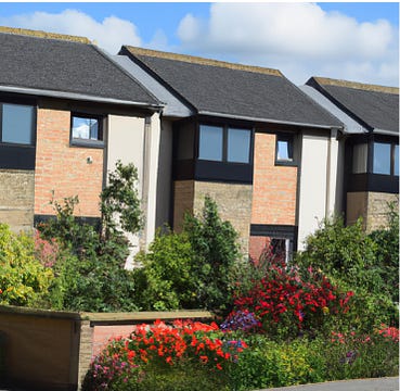 3 2 storey houses with dark window frames and 3 different brick walls. There is mature shrubbery in the front garden and a brick wall surrounding the gardens.