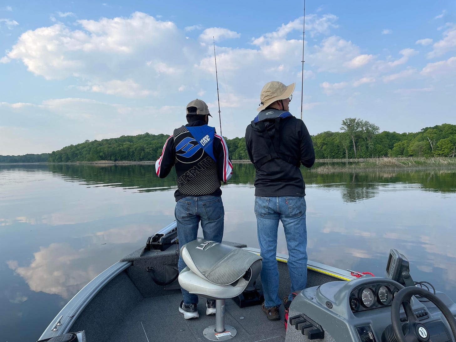 Max and Jon, wearing blue jeans, sweatshirts, life vests and hats stand in the bow of a small boat on a calm lake. The blue sky and fluffy clouds are reflected in the water, and in the distance is a green tree-lined shore. They each hold a fishing rod and are intently watching and waiting for fish to bite.