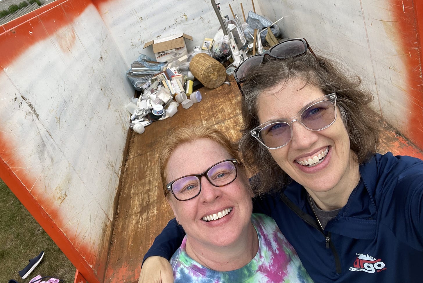 Two women stand in front of a giant Dumpster containing a bit of trash.