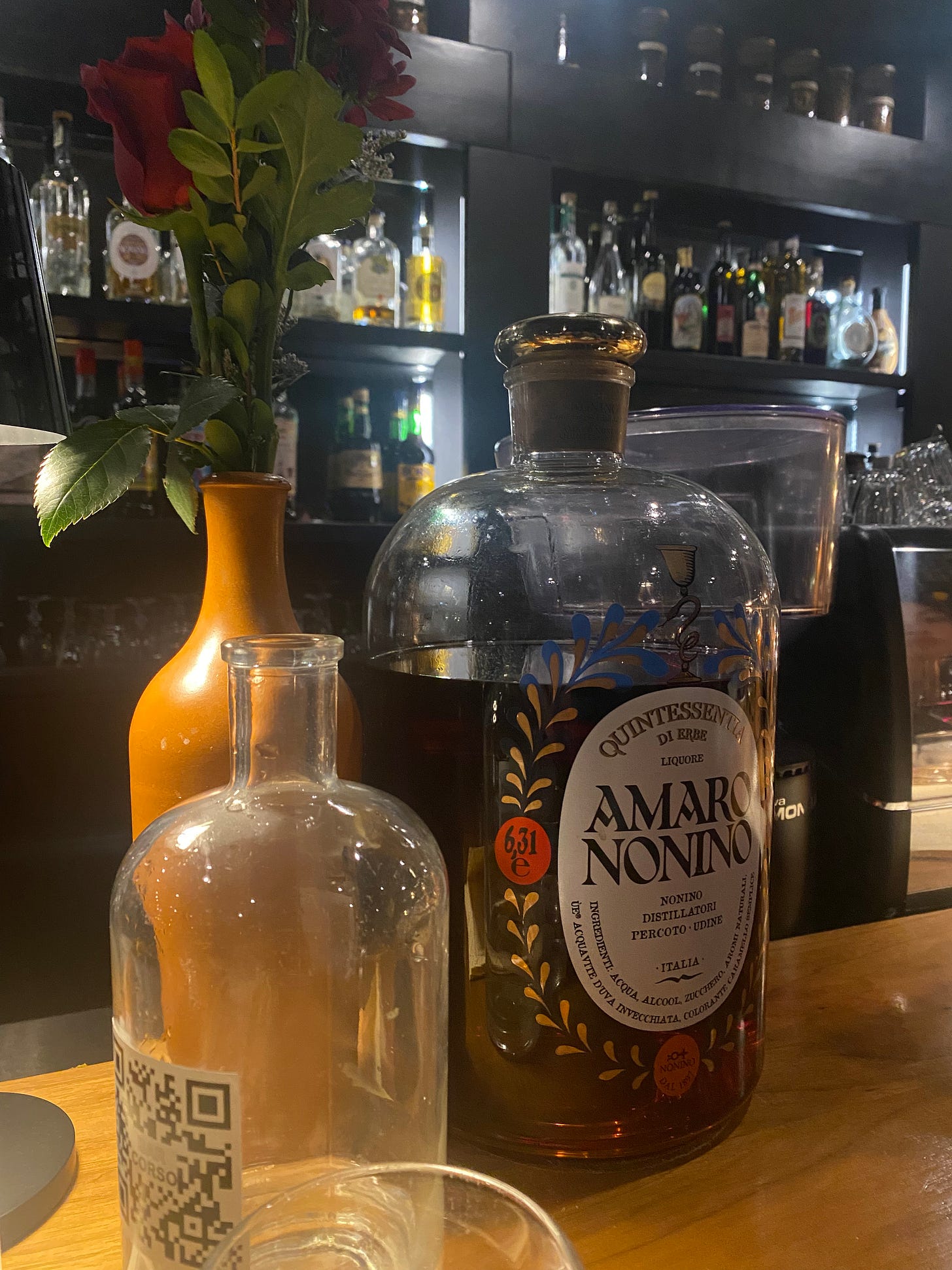 A bar at night, with a painted wine bottle holding a rose with leaves, next to an almost comically large bottle of amaro.