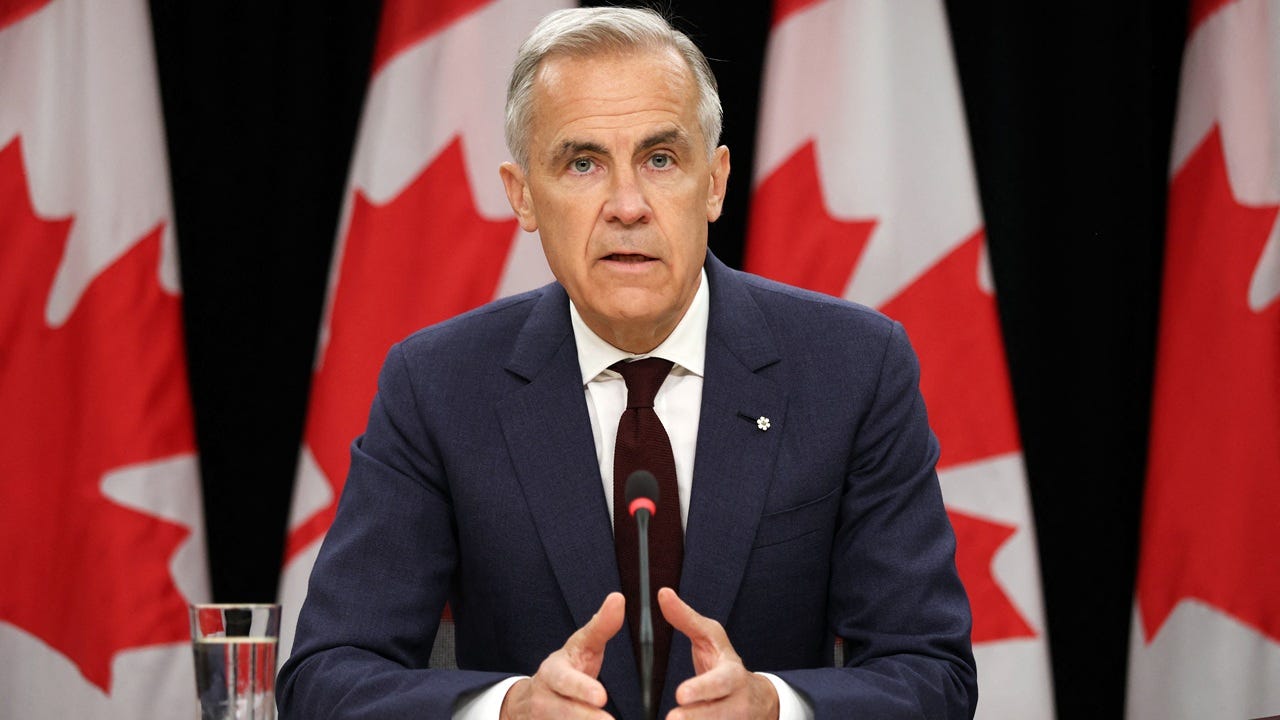 A man in a dark suit speaks at a press conference, seated at a table with a microphone and a glass of water. Several Canadian flags are displayed in the background.
