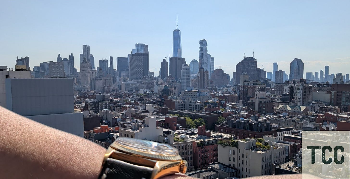 Wrist-shot of the Rolex Day-Date on a green strap, arm resting on a railing, New York City skyline in background.