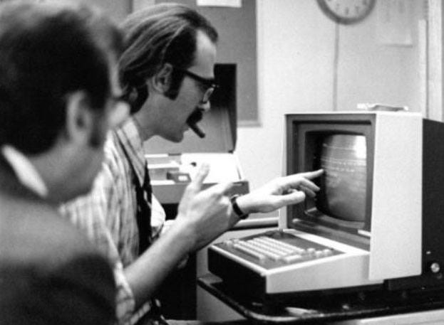A white man touches the screen of an early data entry terminal while another looks on.