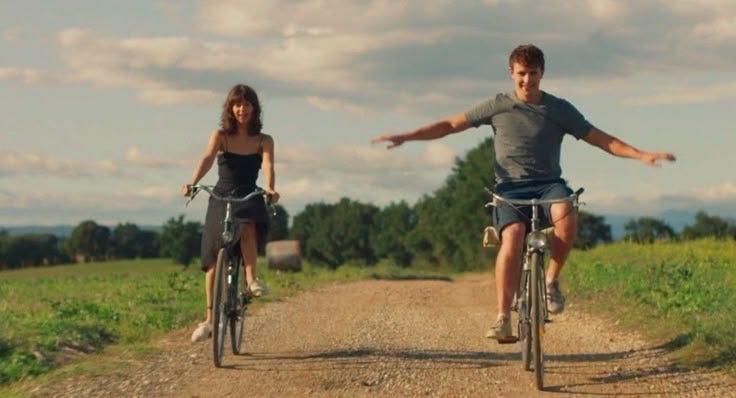 This may contain: a man and woman riding bikes down a dirt road next to a lush green field