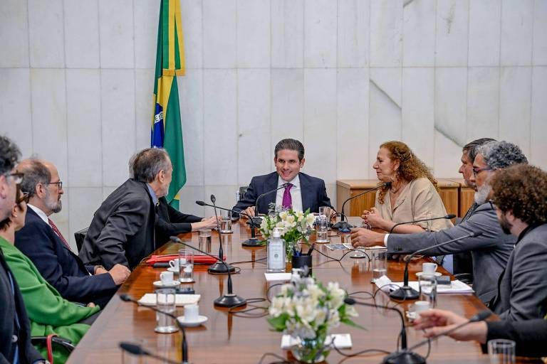 A imagem mostra uma reunião em um ambiente formal, com uma mesa retangular onde estão sentadas várias pessoas. No centro, um homem de terno e gravata está conversando com os demais participantes. Ao fundo, há uma bandeira do Brasil. As pessoas estão vestidas de maneira formal, e há flores em um vaso sobre a mesa. O ambiente parece ser um escritório ou sala de reuniões.