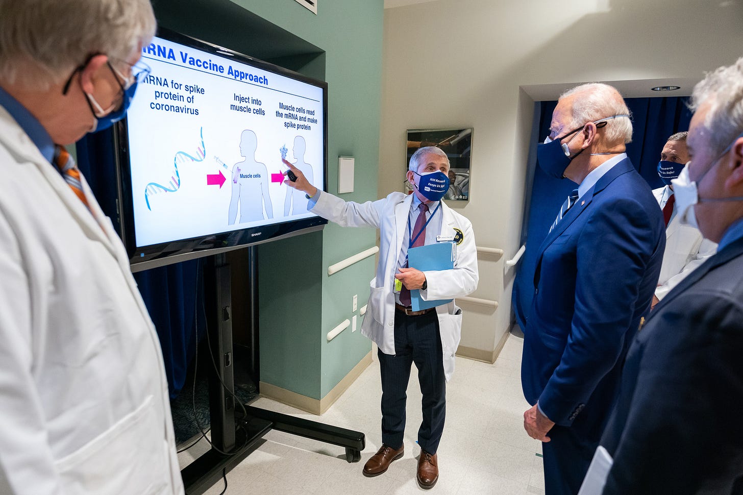 Dr. Anthony Fauci, in lab coat and mask, points to an mRNA vaccine diagram on a screen as President Biden and others look on.