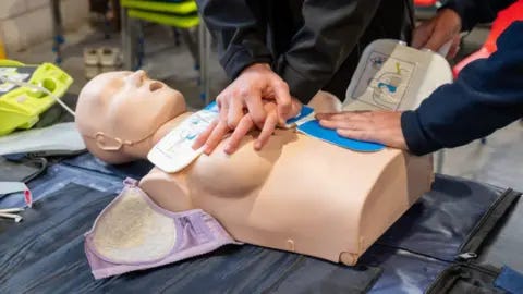 Northamptonshire Police, Fire and Crime Commissioner A female CPR dummy, on a surface, with a bra around it, and three hands on it, there is equipment all around it. You can see three arms. 