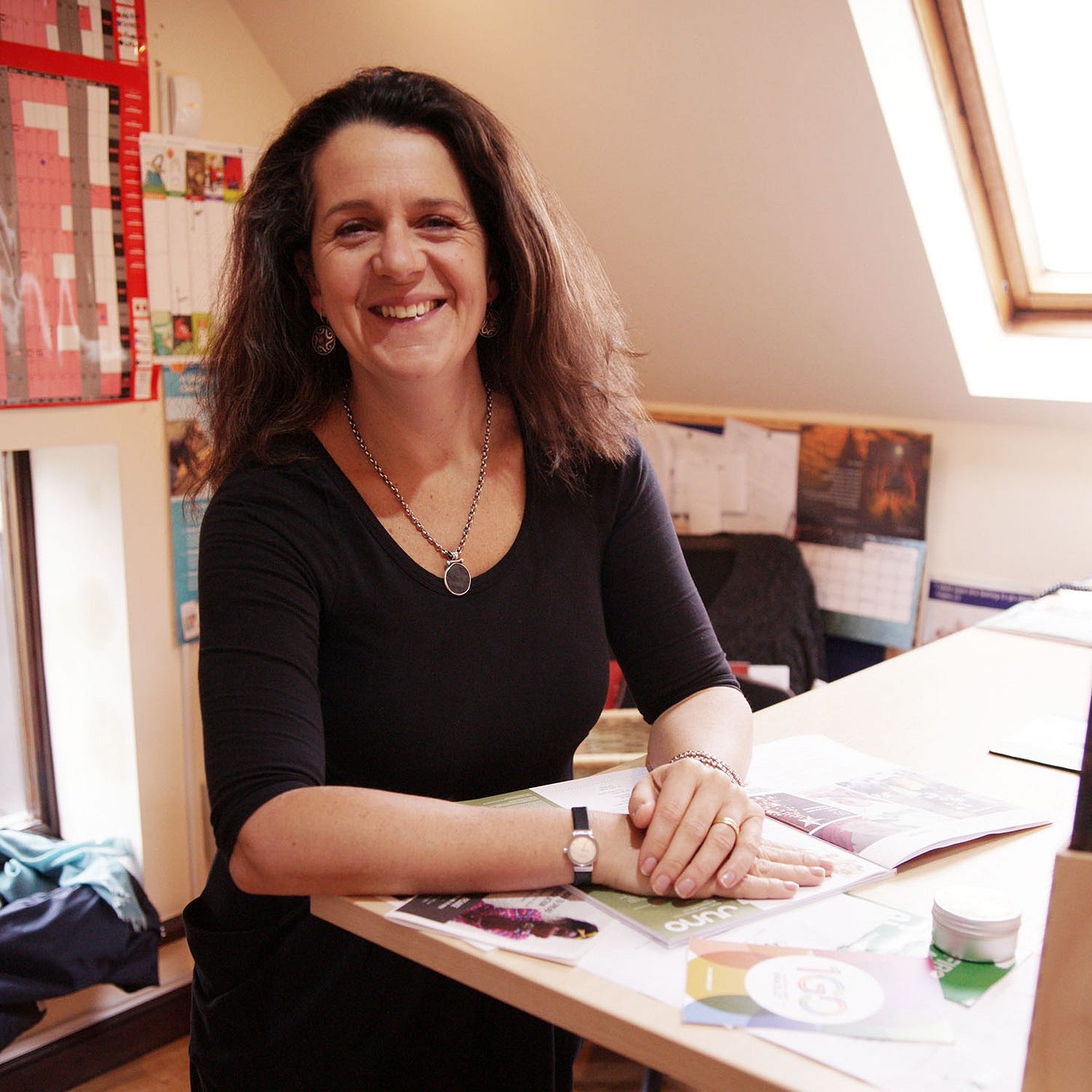 Editor Saffia in a black dress at a standing desk with books, wearing a watch and necklace. Editor Saffia in a black dress at a standing desk with books, wearing a watch and necklace.
