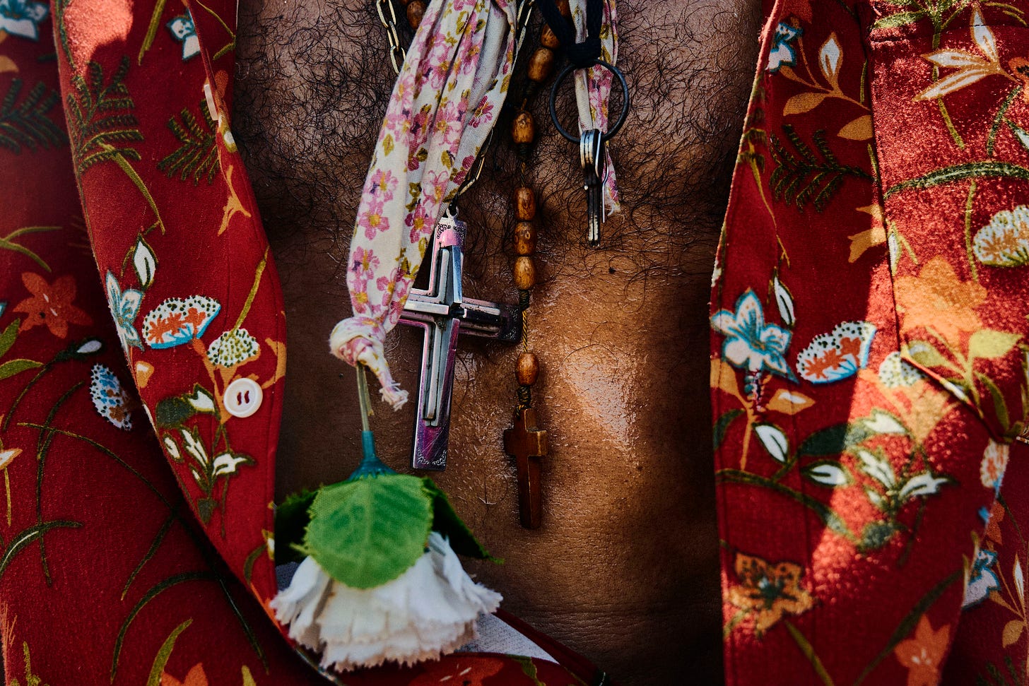 A close-up of a person wearing a red floral shirt, showing layered necklaces with crosses, a wooden bead rosary, a floral scarf, and a white artificial flower resting on their chest.