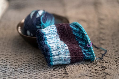 A photograph of a knitting project in process. The yarn is blue, white, and purple against a gray blanket. A photograph of a knitting project in process. The yarn is blue, white, and purple against a gray blanket.