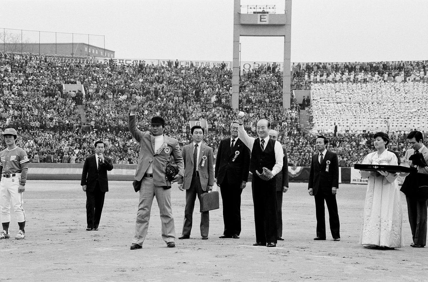 President Chun Doo-hwan, dressed in a vest and tie, prepares to throw a baseball on the pitcher’s mound during the ceremonial opening of South Korea’s first professional baseball game in 1982. Surrounded by officials and players in front of a packed stadium crowd.