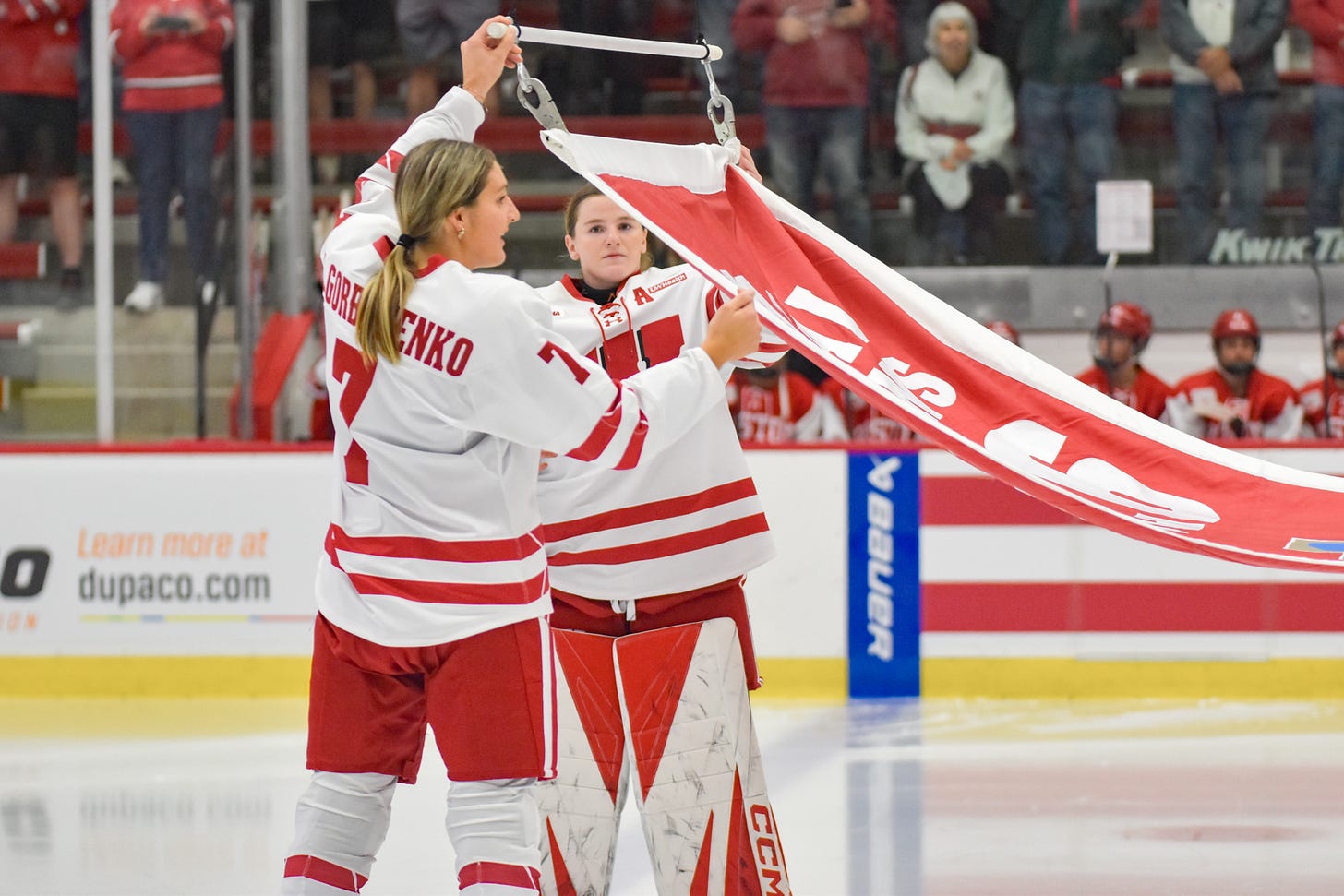 Kelly Gorbatenko and Ava McNaughton connect the Wisconsin women's hockey national championship banner to a set of clips for it to be raised