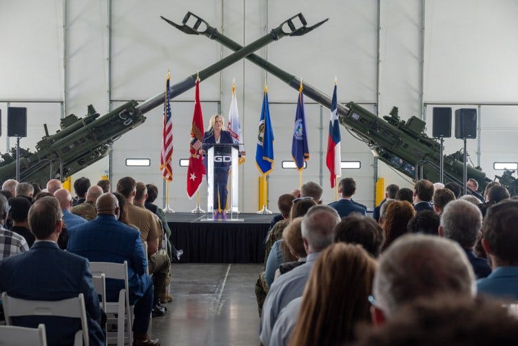 A woman addresses a crowd from a podium branded with the General Dynamics logo, on a stage displaying various flags and two howitzers. A woman addresses a crowd from a podium branded with the General Dynamics logo, on a stage displaying various flags and two howitzers.