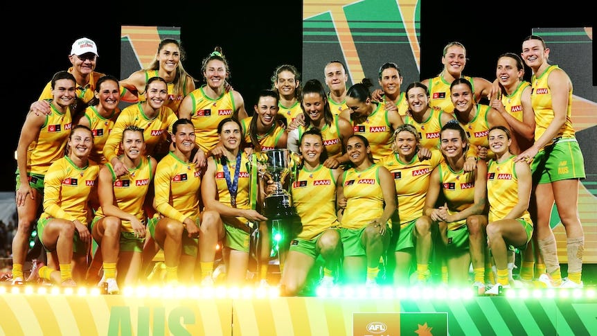A big group of Australian AFLW players gather together to celebrate with a trophy after beating Ireland. A big group of Australian AFLW players gather together to celebrate with a trophy after beating Ireland.