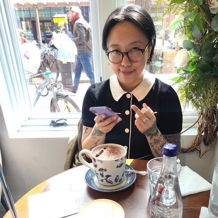 Two images side by side, on the left is K wearing a cute black and white short-sleeved shirt, sitting at a table in a coffee shop in front of a window, giving the camera the middle finger with a cheeky expression. On the right is the same scene, but K is smiling down at their phone.