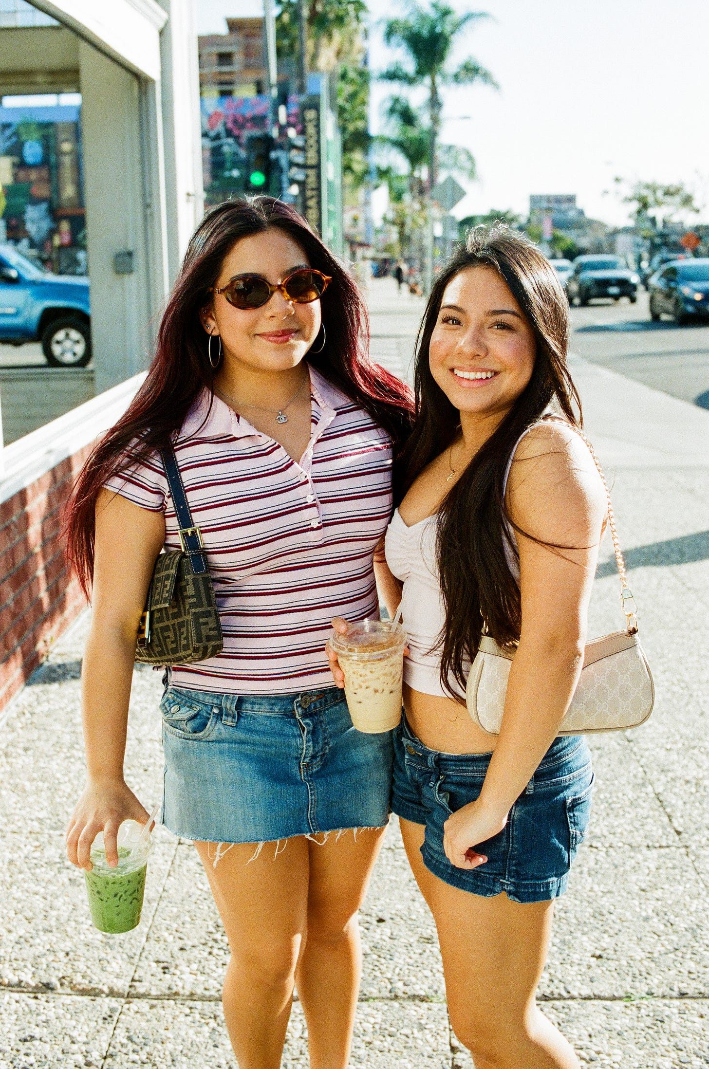 Two women standing on the sidewalk smiling. Each holding a matcha and a coffee. Palm trees in the back and one womans red dyed hair and shirt matching the brick on the building on the left. 