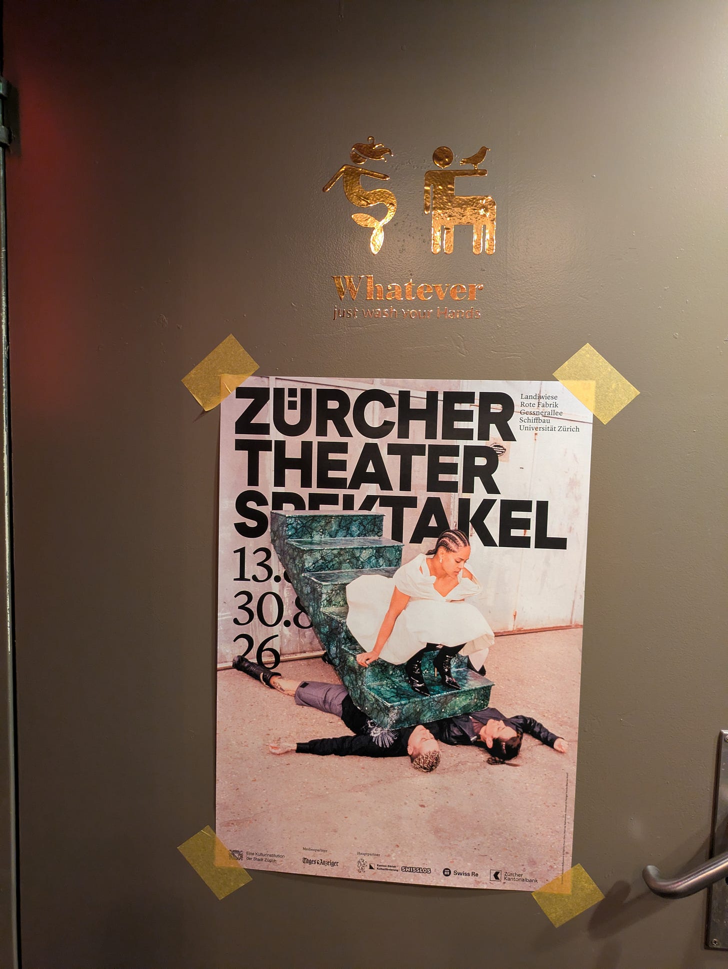 A Zürcher Theater Spektakel festival poster taped to a restroom door beneath a gold sign reading, “Whatever — just wash your hands.” A Zürcher Theater Spektakel festival poster taped to a restroom door beneath a gold sign reading, “Whatever — just wash your hands.”