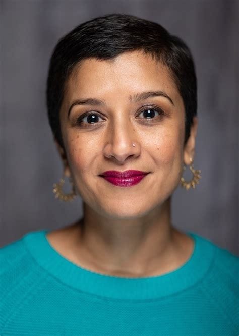 A woman with short dark hair is shown against a softly blurred gray background. She is wearing a teal top, gold hoop earrings, and bright magenta lipstick. Her expression is calm and confident, and she is looking directly at the camera.