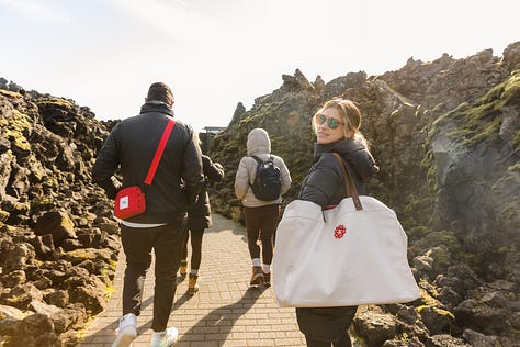 Photo of a coffee cup on a window sill with a view of the outside: green and yellow grass covering rolling hills and gray houses in the middle ground and towering mountain with jagged peaks in the far ground; photo of group of people from behind in winter wear walking along a black sand beach with a tall dark rocky mountainside to the left and a jagged rocky cone island in the background; photo of a black sand beach with large smooth gray and black pebbles in the foreground and disappearing into the horizon with a setting sun; photo of a group of people walking on a stone path with jagged black rocks walling both sides and one woman looking back at the camera; photo of a man and woman smiling posing at a lookout point with large waterfalls in the background below; photo of a woman smiling and holding up a phone, standing on a painted hopscotch on the ground in a bustling pedestrian-only city street; photo of a caramel colored cow being pet on its nose, looking at the camera, and resting its chin on the back of the neck of another cow; photo of a man looking down at his phone as he rests one foot on a higher step of a wall of rocky Basalt columns; photo of several sheep grazing on a field of green grass with a few white buildings in the background and rolling green and yellow-green hills in the distant background.