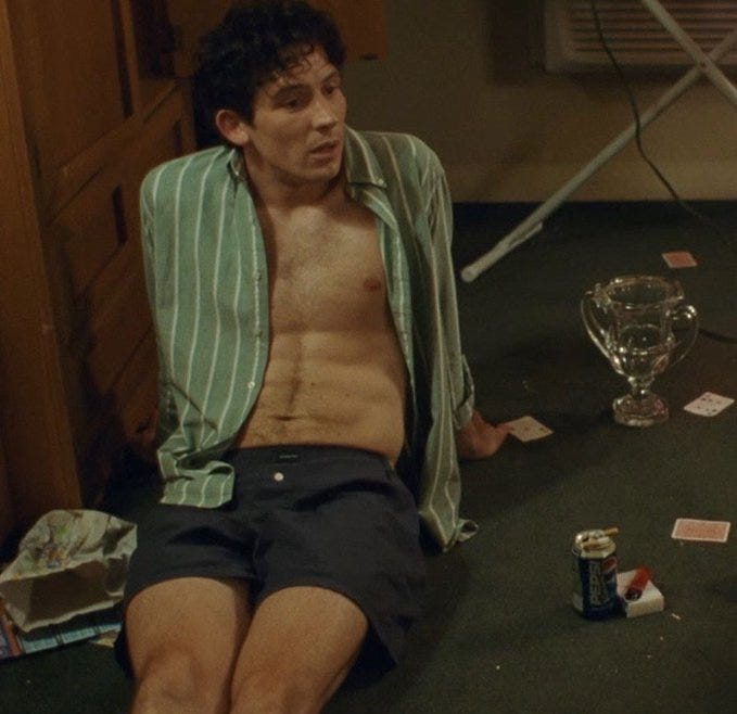 Still from the movie Challengers showing character Patrick sitting on the floor wearing an open green Oxford shirt with white vertical stripes